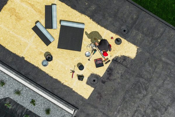 A worker repairs a roof in a sunny environment using various tools and materials. The roof shows sections being covered while others remain bare.